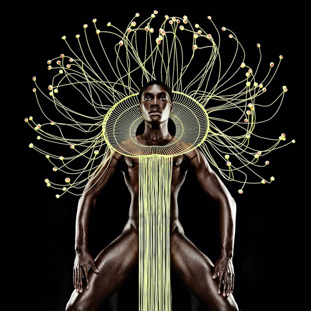 A Black man stands in front of a stark black background. He stands with his legs apart, fingertips resting just above his gently bent knees. He wears a golden collar with tendrils radiating around his head and trailing down his bare torso.