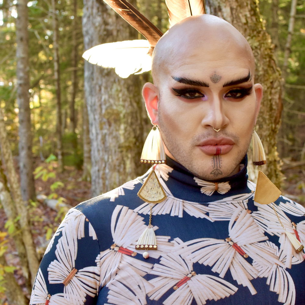 Geo, a light-skinned indigenous person, looks into the camera. Traditional markings are tattooed on their forehead and chin, and their shaved head is adorned with feathers. They have a light beard and dramatic makeup, and are wearing earrings made from tusk shells and porcupine quills. Their clothing is decorated in a tusk shell butterfly pattern.
