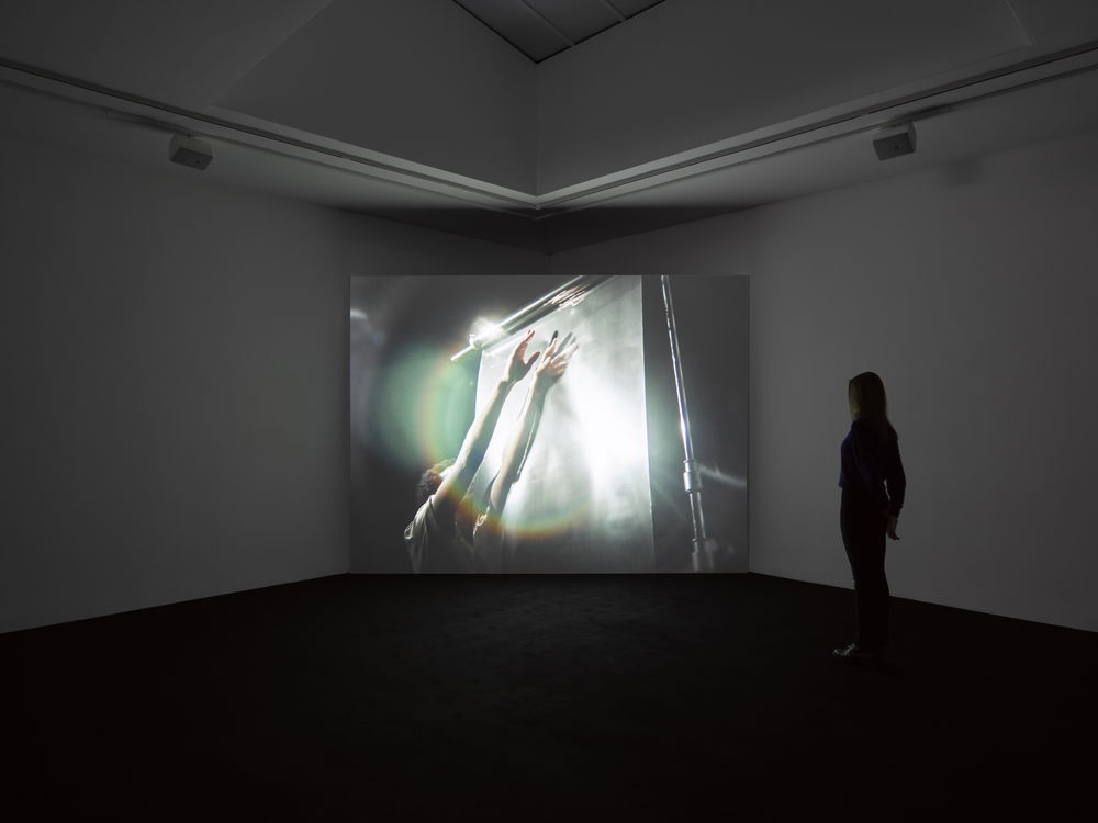 A dark gallery where one person stands facing a small screen in the corner of the room. The screen shows an image of a person seen from the waist up as they raise their hands outward and upward in front of a scrim. The scrim reflects a bright, silvery light and creates a rainbow halo around the person's outstretched arms.