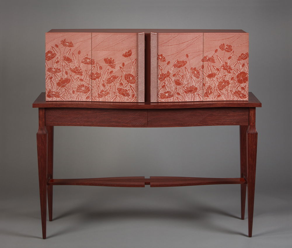 A reddish-orange four-doored cabinet with a poppy landscape carved on its surface sits on a coffee-colored long, slender table. Both the cabinet and table have a serpentine front and textured line designs on their surfaces.