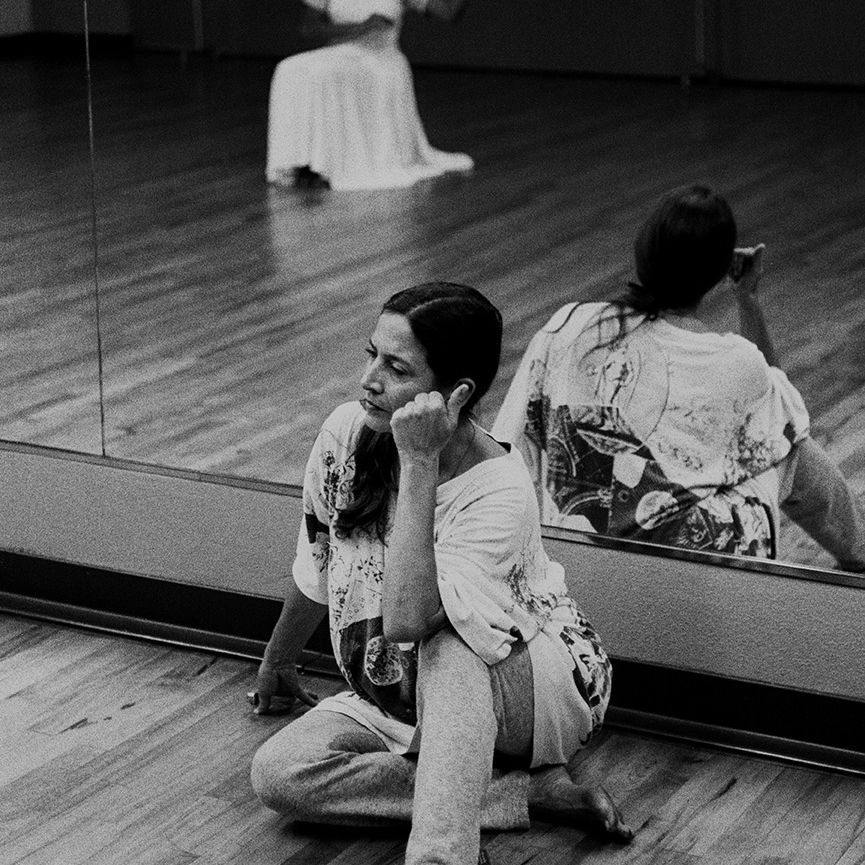 Fotografía en blanco y negro de Petra Bravo y la bailarina Dolores Pedro. Petra está sentada en el suelo contra el espejo del estudio de danza, mirando pensativa a Dolores con la cabeza apoyada en los nudillos. El reflejo de Dolores en el espejo la muestra arrodillada con un vestido blanco.