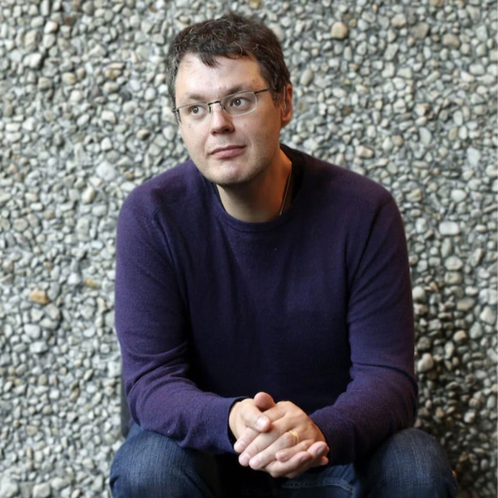 A person wearing rimless glasses and a purple sweater sits in front of a stone wall looking slightly away from the camera.