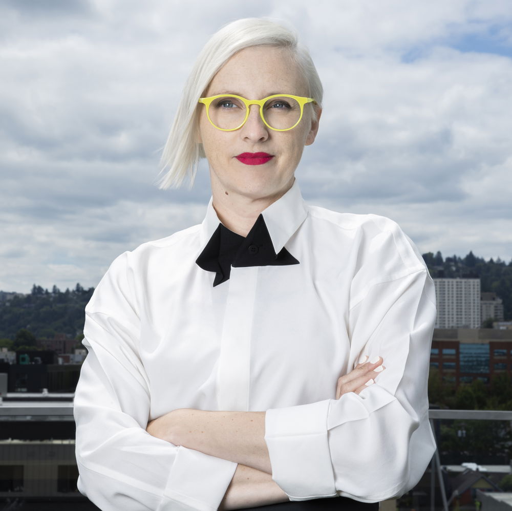 Jennifer, a woman with blonde hair, stands with arms crossed wearing lemon-yellow glasses and a fake tuxedo shirt.