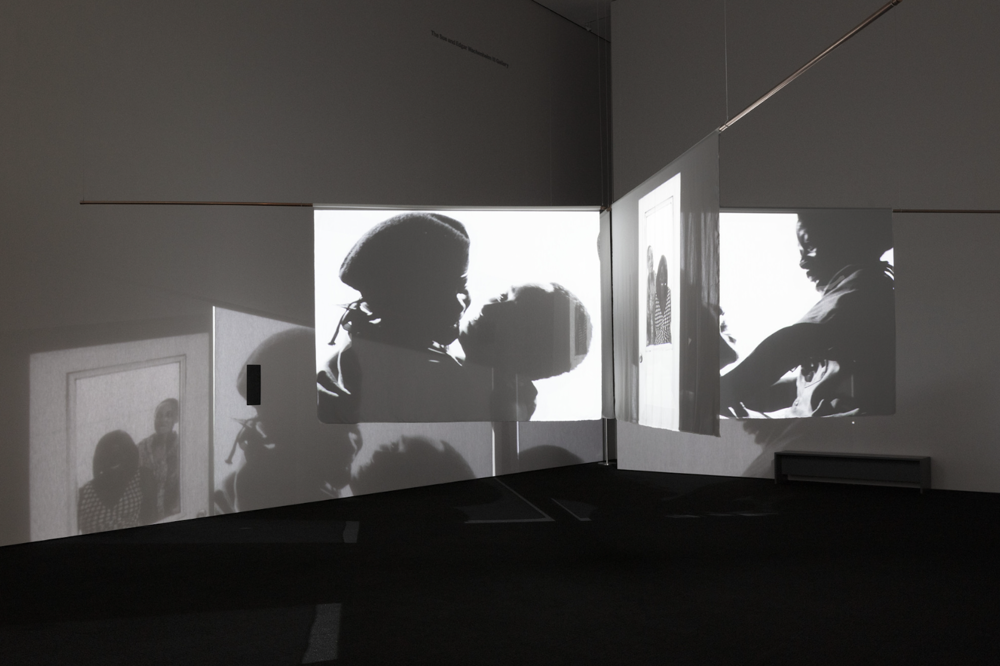A dark gallery with three screens adjoining perpendicularly; two run on a horizontal axis, and one intersects in the middle. Each has a different image of a Black couple in different poses. The images project off the screens and onto the empty walls, creating a dizzying effect.