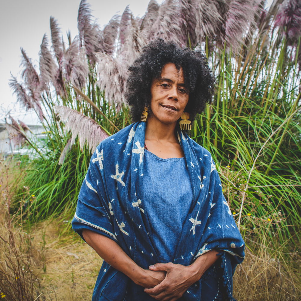 Amara, a Black woman with dark hair, stands in front of tall grasses blowing in the wind and smiles gently. She wears an indigo shawl and dangling wooden earrings.