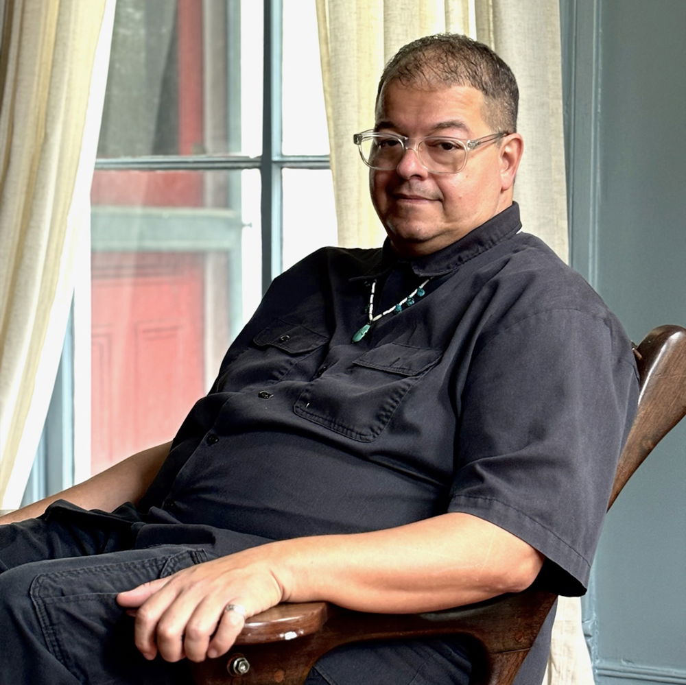 A man with close-cropped hair and clear-rimmed glasses sits in a wooden chair by a window, gazing out at the viewer. He is wearing a turquoise necklace and all-black clothing.
