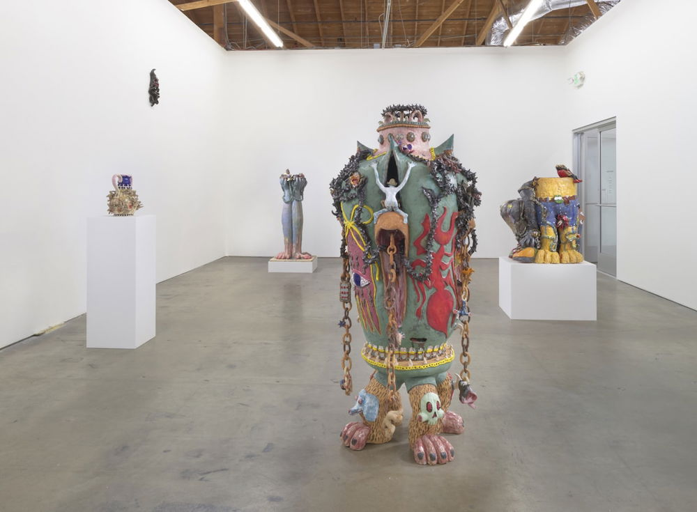 An in-gallery photograph, displaying several sculptures. The centralized sculpture is a human-sized vessel balanced on three hairy legs resembling that of a primate. The vessel itself, huge and urn-like, is green with graphic images painted on its sides. It is covered in chains, barbed rope, and small figures.