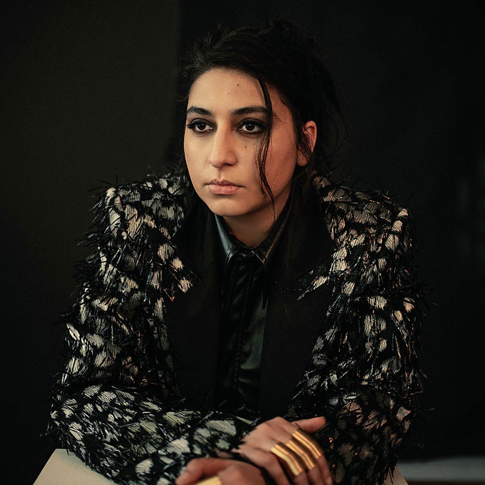 A Pakistani woman poses against a black background, leaning over a table with crossed arms. She wears a black and gray blazer with gold rings on her fingers and looks to the left of the camera.