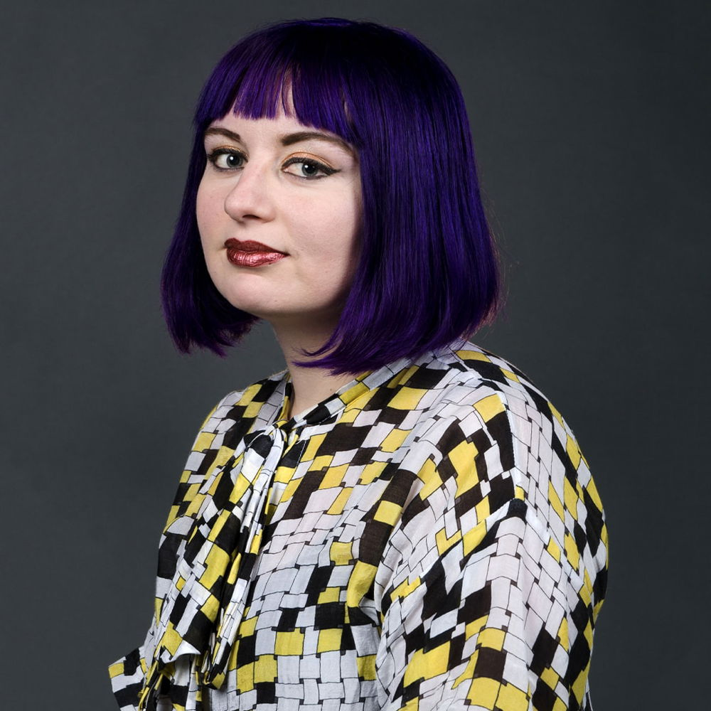 Headshot of a cisgender woman with short purple hair wearing a yellow, black, and white tile-patterned shirt. The woman has pink tinted skin and dark red lips with a high gloss finish.
