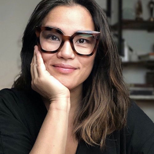 A portrait of a Filipino American with dark brown hair and large tortoiseshell glasses. She looks directly at the viewer, smiling, and resting her head in her right hand.