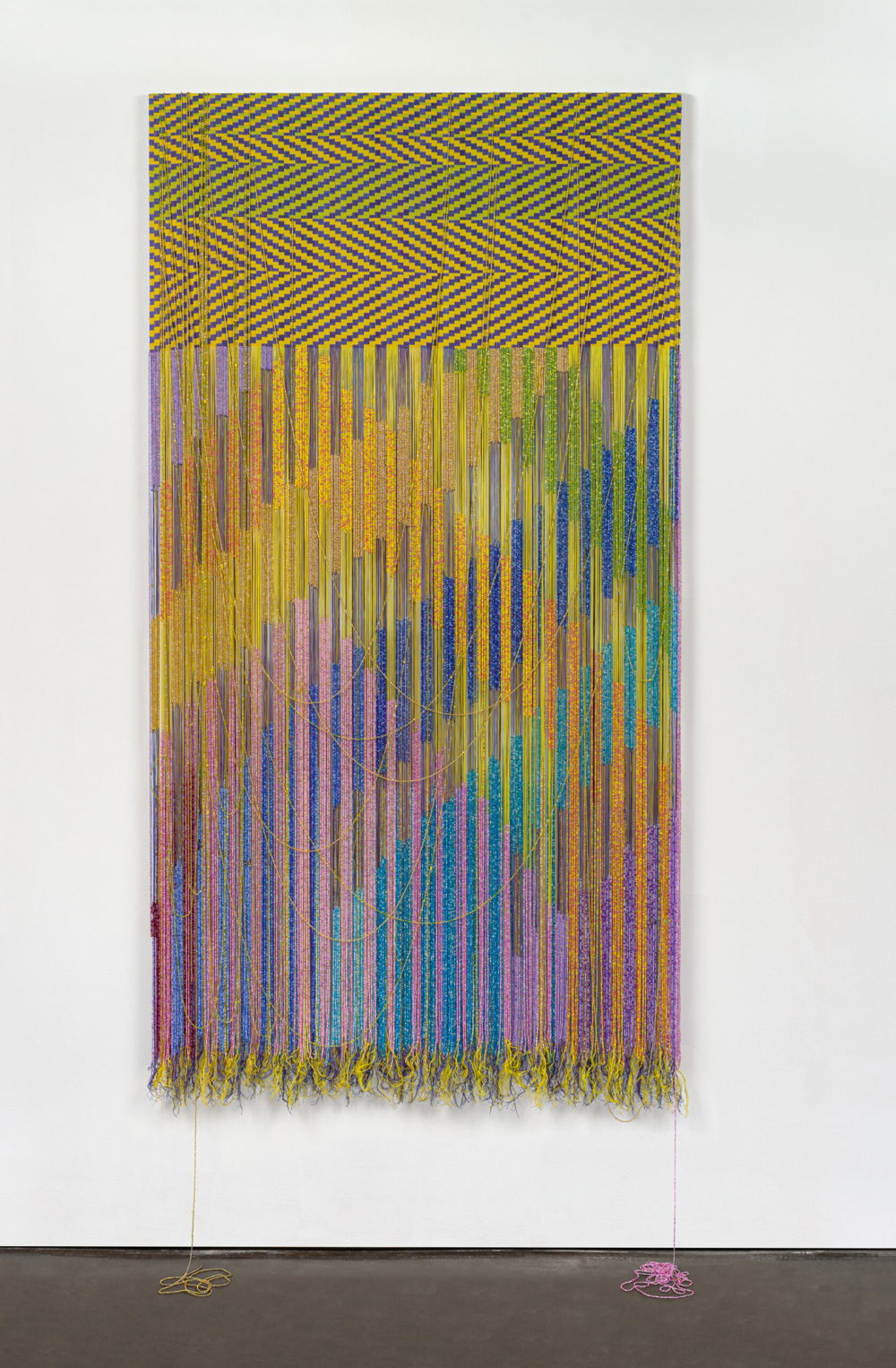 A vertical rectangular-shaped tapestry. The top quarter of the composition is a completed tapestry composed of geometric zig-zag patterning in electric yellows, greens, and violets. The bottom three quarters of the composition are unwoven with intricately-beaded strands of thread that create a vibrating pattern of criss-crossed diagonals in glowing yellows, oranges, pinks, greens, blues, turquoises, and purples. A few loose strands are wisped upward creating thin U-shaped lines. Additional loose strands dangle downward to puddle onto the floor.