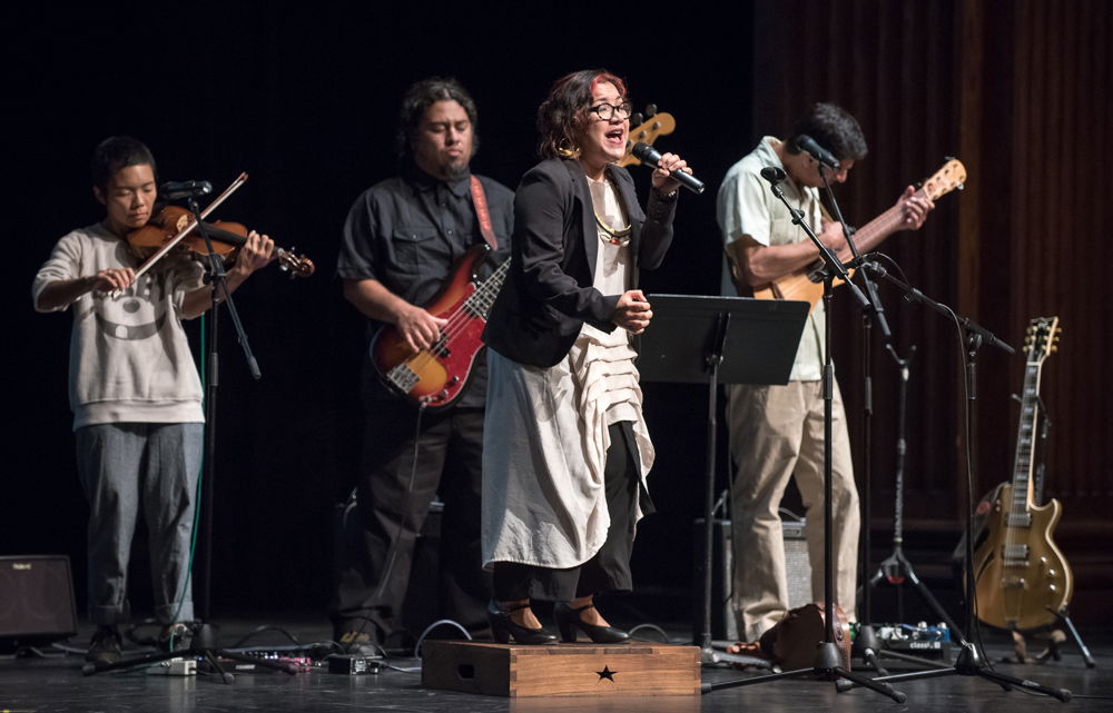 Martha performs with a band onstage. She stands on a small podium and sings into a microphone, while musicians behind her play various string instruments.