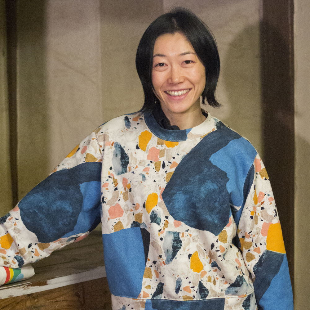 Aki, a Japanese woman with medium length black hair, stands by an empty shelf space in an old Japanese apartment building. She has a big smile and is wearing a loose sweatshirt with bold colorful prints.