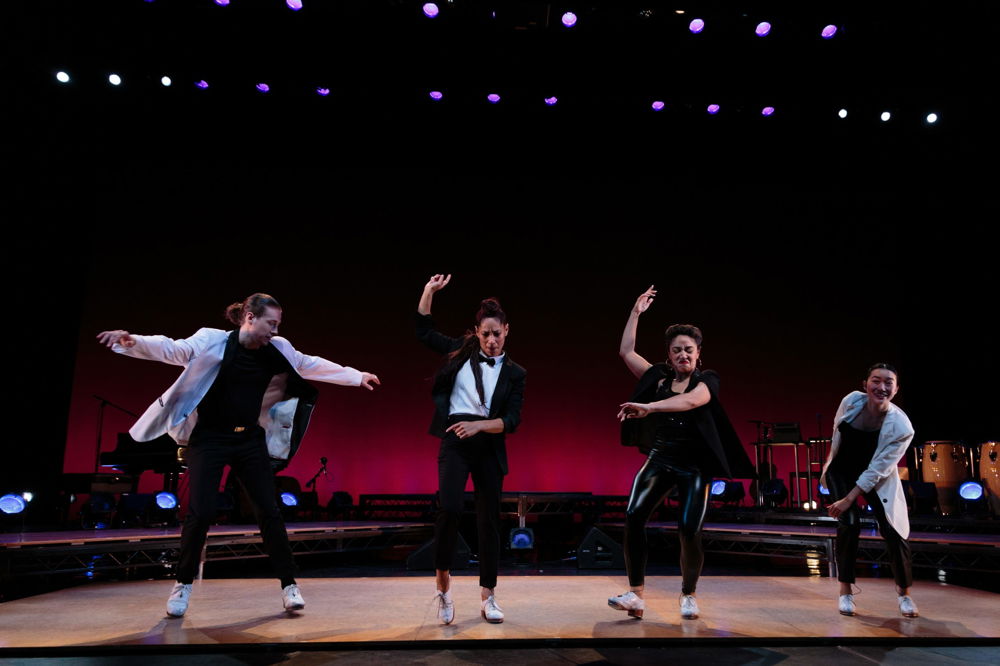 Four dancers mid-motion on a swanky stage with a maroon curtain backdrop. Their motions are energetic; their black-and-white costumes are elegant and fun.