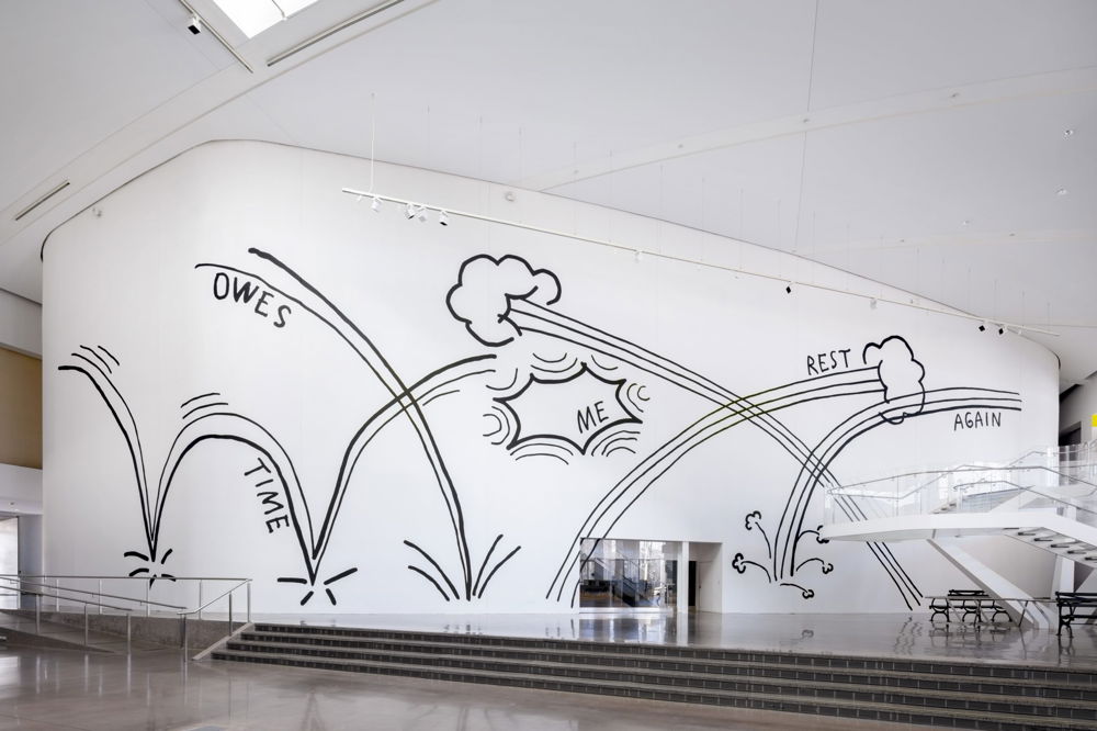 Image of a large-scale artwork covering the interior white wall of a building. The words “Time Owes Me Rest Again” are written in black against the wall, given motion by sweeping comic book style lines that imply the words are bouncing across the wall or being hurled from the sky.