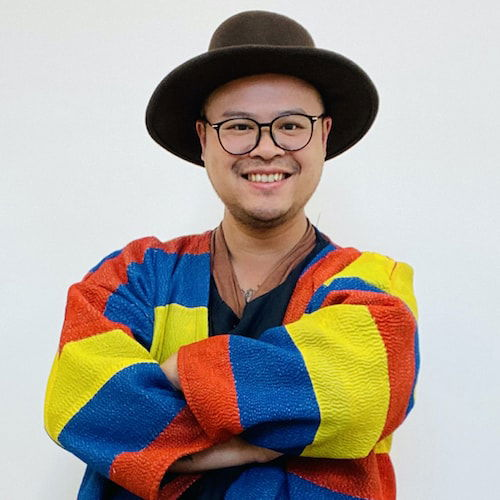 A portrait of a Chinese/Filipino individual standing in front of a white wall. They smile warmly at the viewer with their arms casually crossed at the chest. They wear a wonderfully bright colored, quilted jacket; striped red, yellow, and blue.
