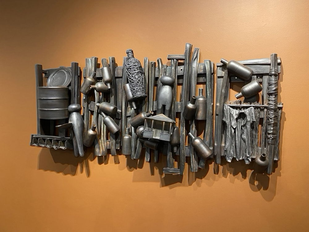A metallic sculpture hung on a yellow wall. The sculpture resembles a jumble of objects on top of wooden pallets. Objects include bottles, giant clothes pins, a large tin can, a miniature house, and a button-down shirt.