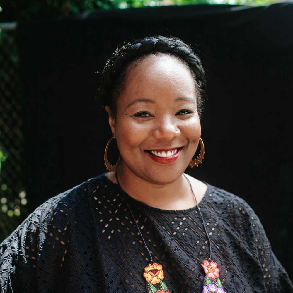 Maori, a Black woman with a braided updo, stands outdoors against a dark background. She wears a black cotton blouse, an embroidered flower necklace, large brass hoop earrings, and smiles at the camera.