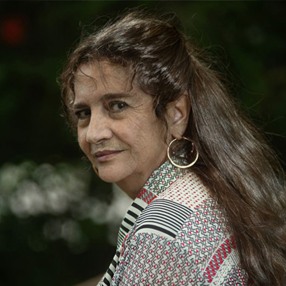 An older woman with long brown loose hair smiles softly at the camera. She is wearing a patterned blouse and hoop earrings.
