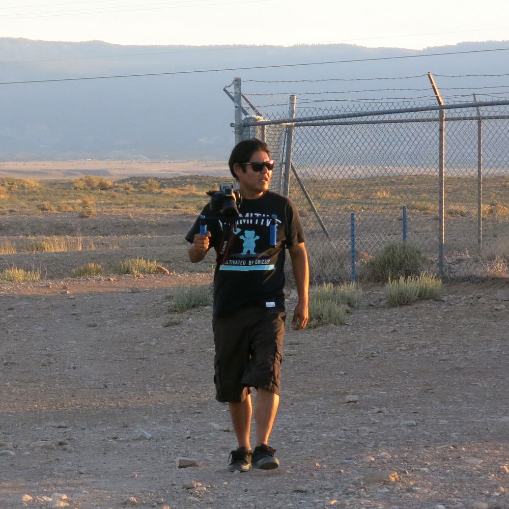 ID: A man dressed in a dark t-shirt, shorts, and sunglasses holds a camera as he walks in a mountainous desert landscape, just in front of a barbed wire enclosure.