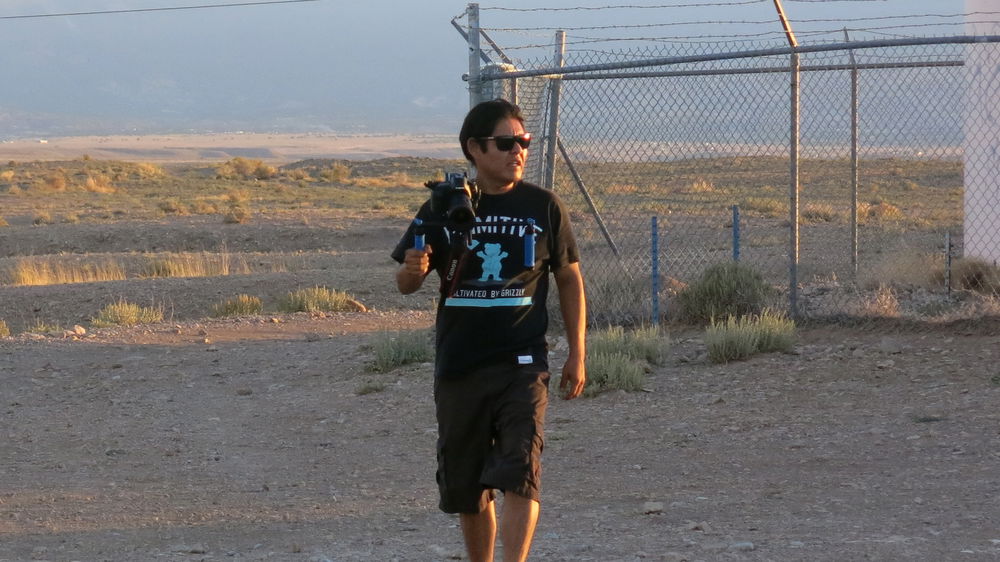 ID: A man dressed in a dark t-shirt, shorts, and sunglasses holds a camera as he walks in a mountainous desert landscape, just in front of a barbed wire enclosure.