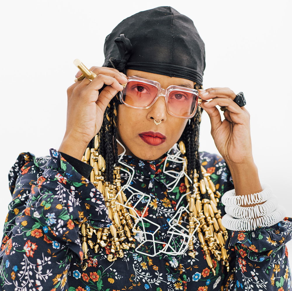 Tiff, a young Black woman, is heavily adorned by the jewelry she created and rocking a du-rag, gold beads, and a floral dress. She holds her glasses as she gazes into the camera.