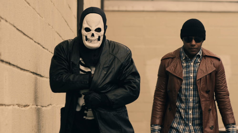 Two men walk toward the camera. The man on the left wears a skull mask, skeleton t-shirt, and a black leather trench coat. The man on the right wears a dark colored beanie on his head, sunglasses, a plaid collared shirt, and a brown trench coat