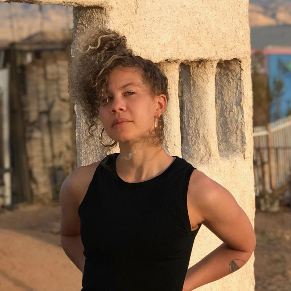 A light-skinned queer Black woman with a blonde ponytail and gold hoop earrings stands in front of an adobe structure in a black tank top with her hands on her hips, facing the camera.