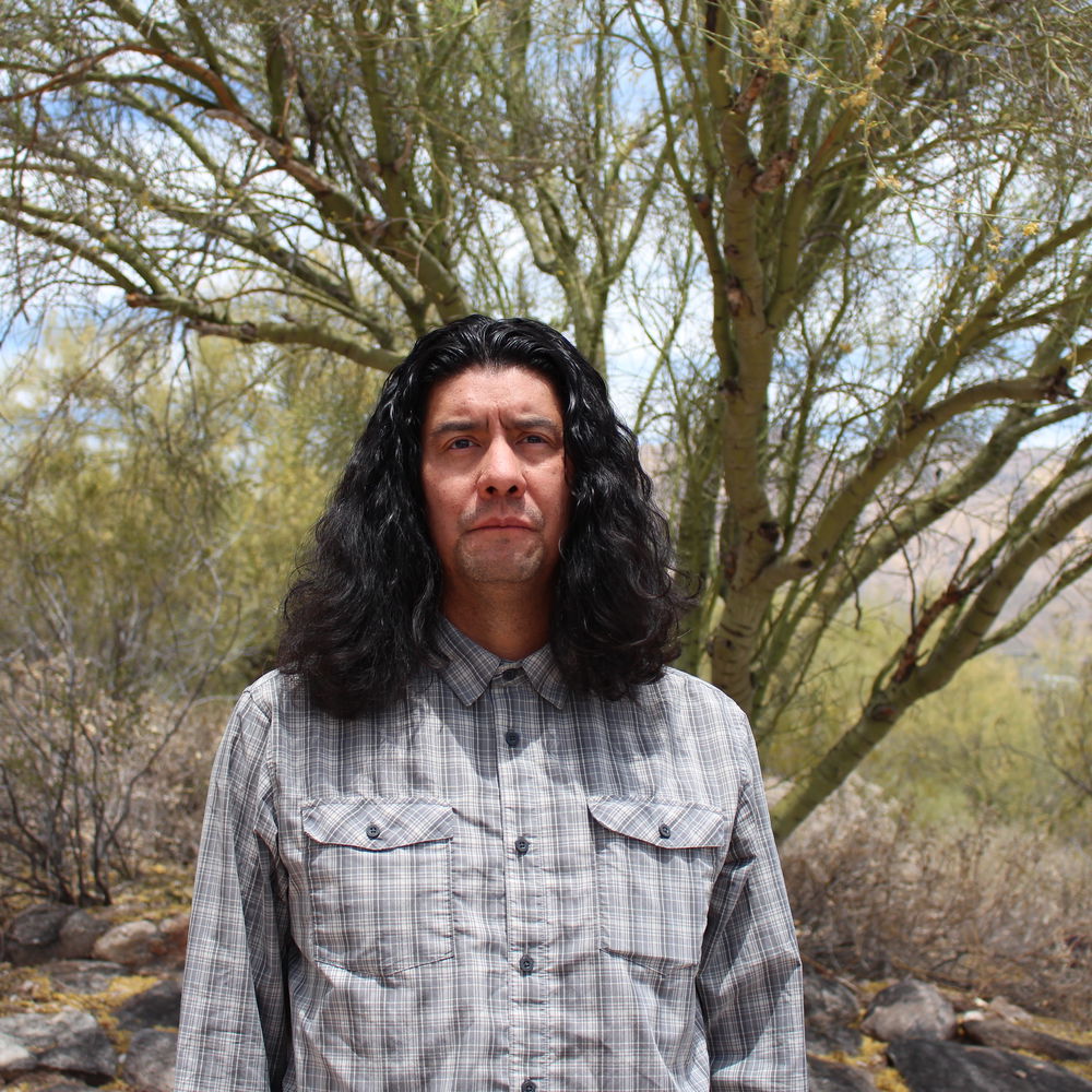 A man with long, wavy dark hair stands in front of a natural landscape with a palo verde tree and other bushes. He looks into the camera with a serious expression.
