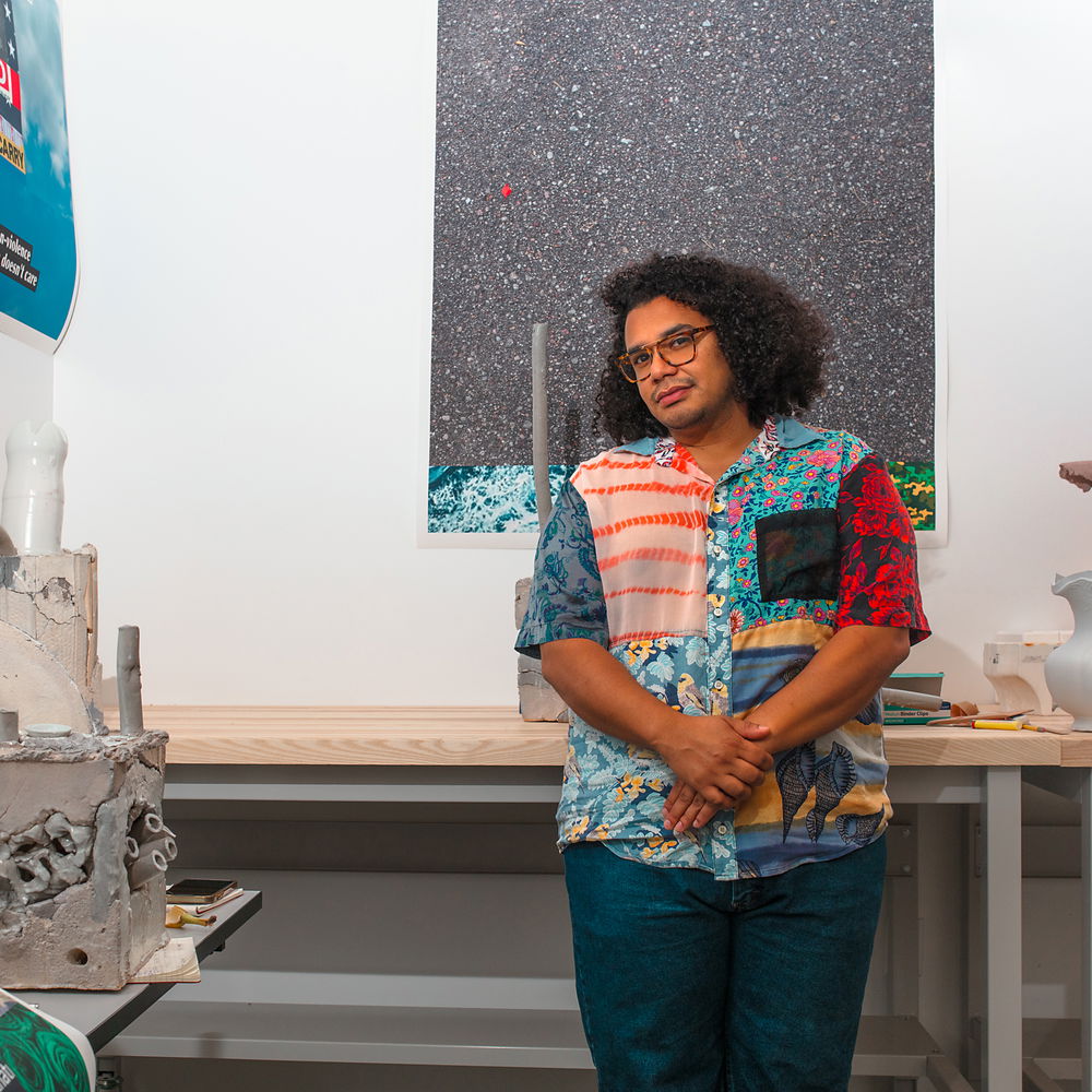 A man standing in a studio space wearing a patchwork silk top made from several different patterned fabrics, turtle-shell glasses, and curly-hair afro. Around him are sculptural works and digital prints of different collages he composed on the computer. 