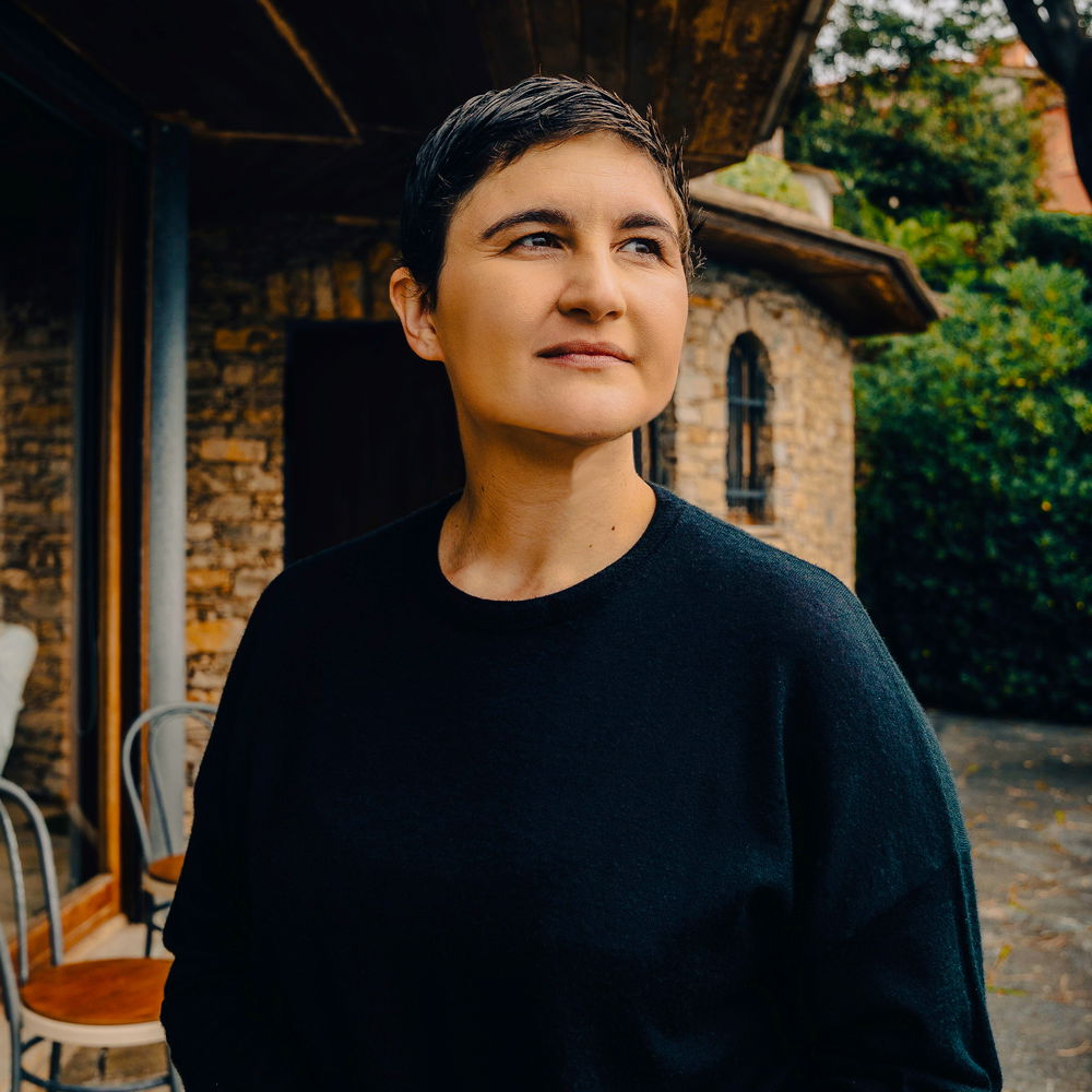 A person with medium-tone skin and short brown hair wears a black sweater on a balcony, looking out at the scenery.