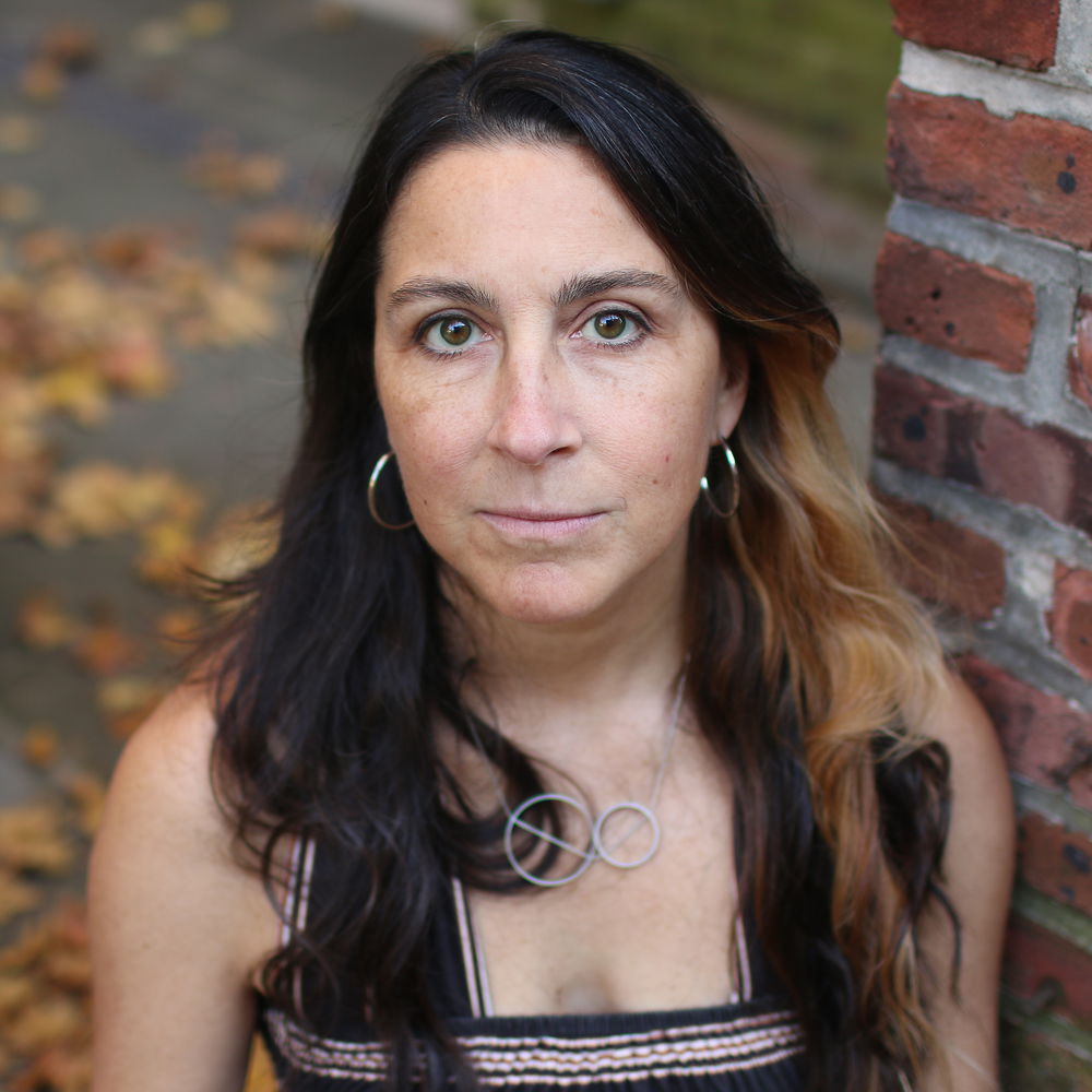 A head shot of a woman, seen from a raised angle. The woman has a light complexion, big brown eyes, and brown hair with a streak of copper in it. She stands with a neutral expression next to a red brick wall on a wet rainy autumn day.