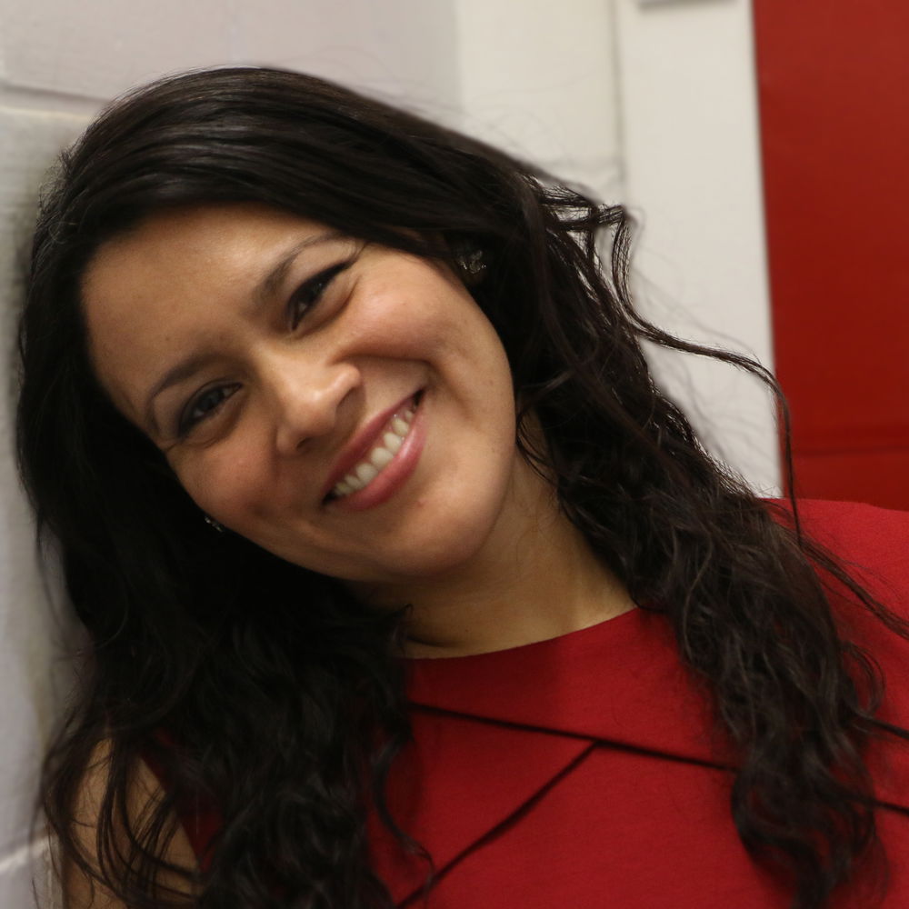 A woman with long black hair beams with a big smile while tilting her head to the left. She poses in the corner where two walls meet. One wall is white and the other is red, matching the red dress she is wearing.