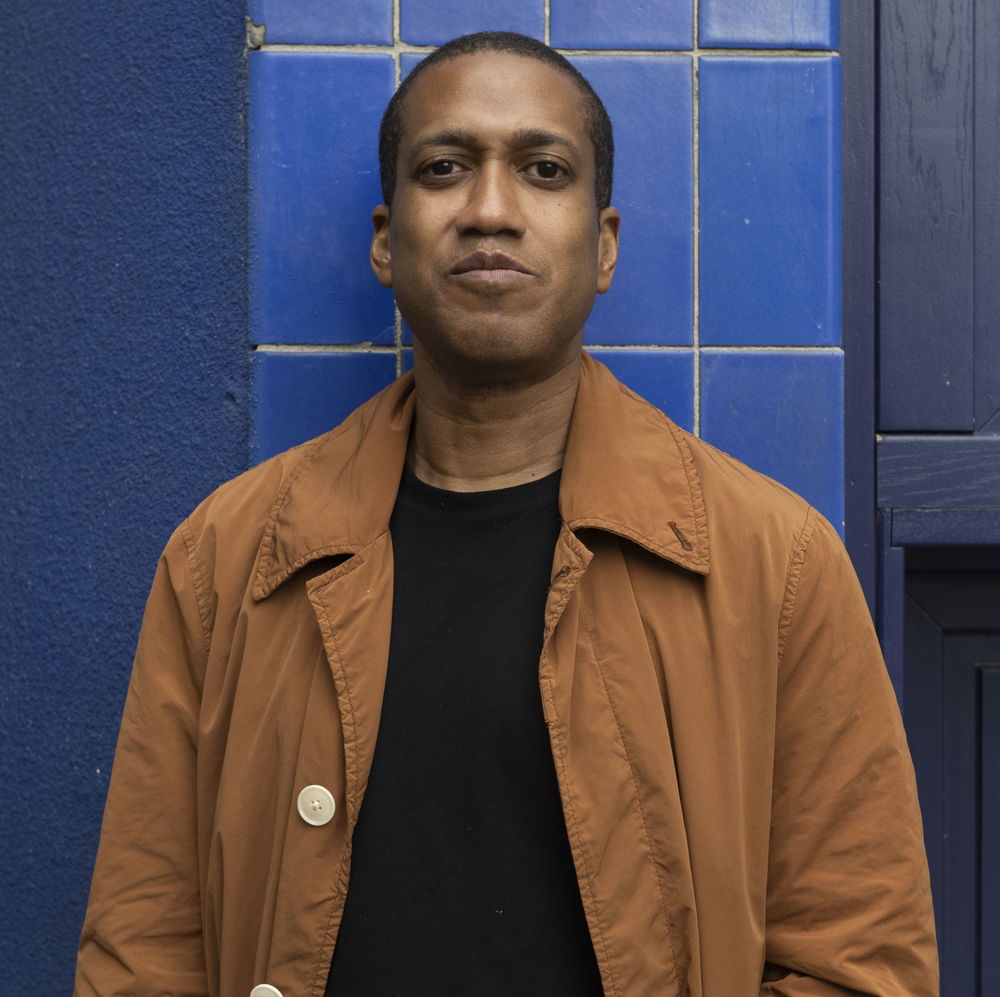 A headshot of a person with a brown complexion and short cropped hair stands in front a blue tiled exterior. He suppresses a little smile and wears a brown coat over a black shirt.