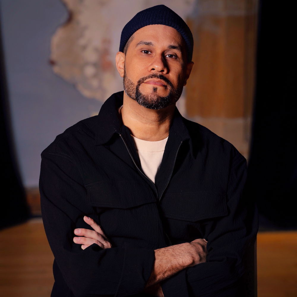 A Latinx man with a short beard wearing a long-sleeved black shirt and a navy blue beanie sitting down with his arms crossed.