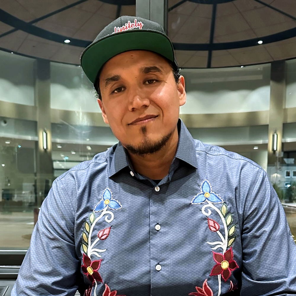 A Native man smiling in front of a large window wearing a black fitted cap and a grayish-blue collared button-up shirt with Ojibwe-style floral applique in blue, yellow, and red. The flowers sway from left to right, as a real flower might. 