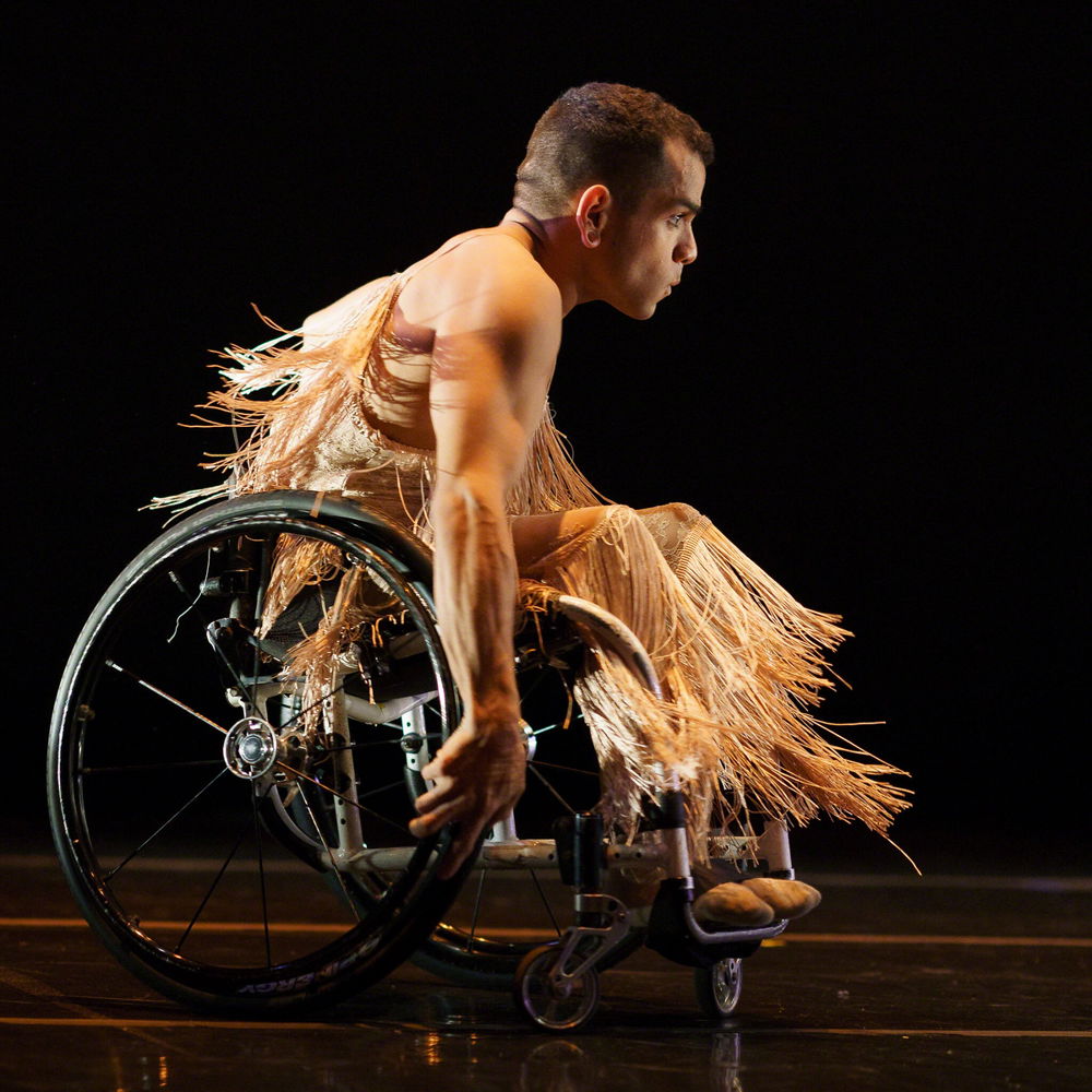 JanpiStar performs on stage in their wheelchair, the strands of their flesh toned, fringed bodysuit swaying with their motion. They have light brown skin, close-cut dark hair, and an athletic build.