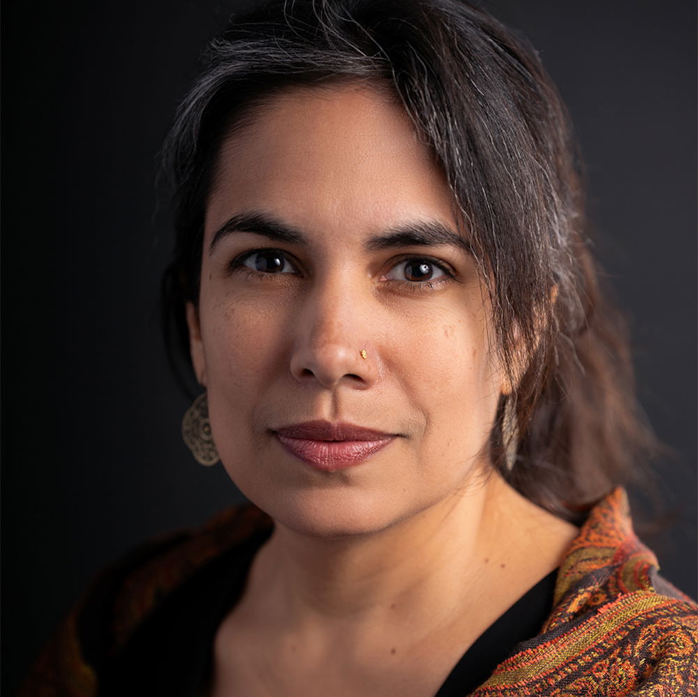 A Punjabi-Chicanx artist with brown skin, dark-brown eyes, and dark, gray-streaked hair pulled up into a loose twist looks into the camera. They are posed against a black background, wearing a black shirt, their grandmother’s orange patterned scarf, earrings, and a nose ring.