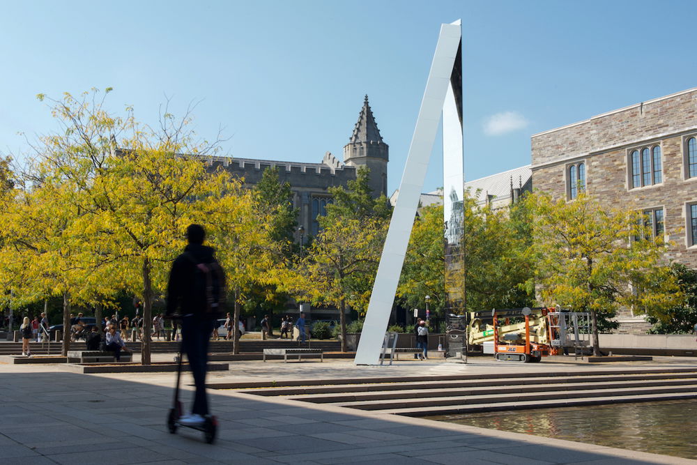 A towering sculpture of two sleek, gray beams leaning against one another in a sunny, verdant plaza. Someone rides by the plaza on a scooter.
