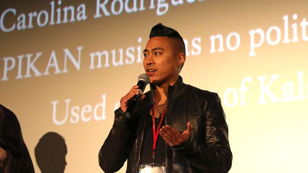 A person of Asian descent with a short mohawk hairstyle, wearing a leather jacket and a red lanyard around their neck with a festival badge attached, speaking into a microphone in front of text projected onto a film screen in the background