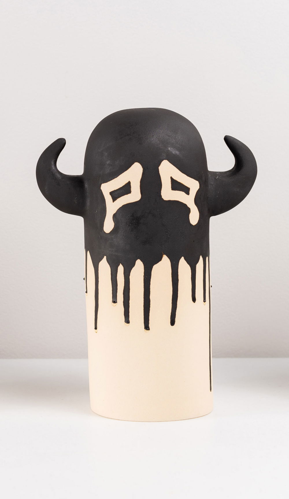 A cylinder shaped sculpture with a rounded top and two antlers protruding on either side. Black paint covers the top half of the otherwise white sculpture, with the exception of two white, cartoonish eyes which drip as though they are crying.