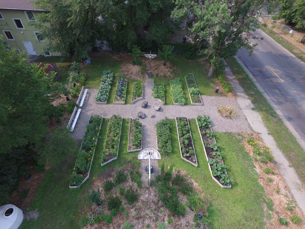 An overhead photograph of a garden formed of twelve raised beds arranged symmetrically to create a large pathway through the center, which contains three decorative rocks. The beds are filled with leafy green vegetables.