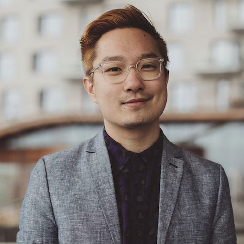A portrait of a queer Chinese American standing in front of a large beige apartment building with many windows and balconies. His hair is dyed an orangey-blonde. He wears clear-framed glasses, a dark purple button-down with gray polka dot pattern, and a gray blazer. His facial expression is gently joyful.
