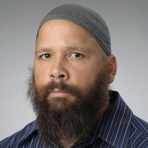A headshot of a man with tan skin and a reddish-brown full beard. His expressionless face is seen from a three-quarter view. He wears a grey kufi, and a navy-blue collared shirt with sky blue and olive green vertical stripes.