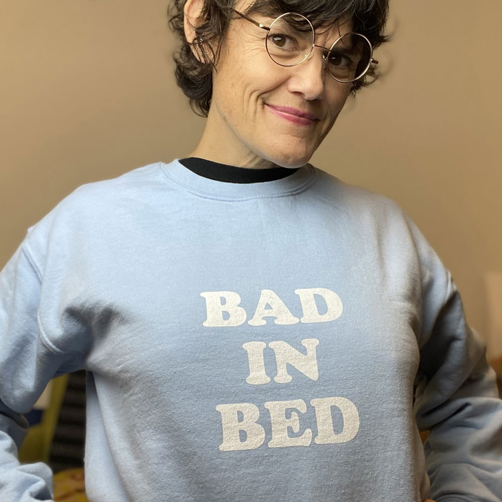 Jibz, a middle-aged queer white woman with brown hair and glasses, wearing a powder blue sweatshirt by the artist Pippa that says “Bad in Bed.”