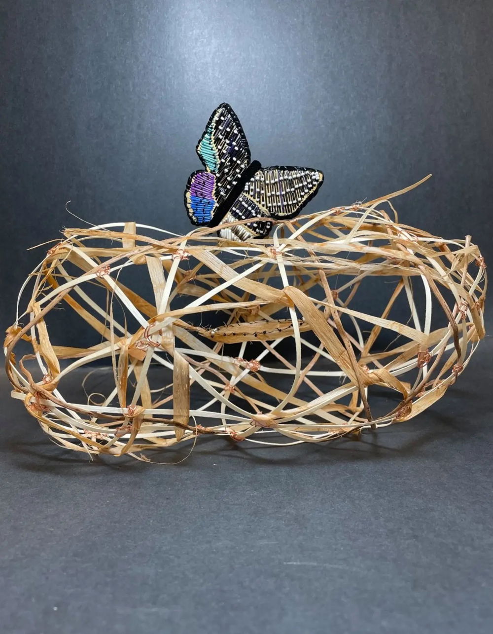 [ID: A small, oblong sculpture constructed from knotted strands of fiber. The fiber resembles that of dried grass: golden beige in color and fragile with fraying edges. Perched on top of the precarious structure is a solitary butterfly with purple, blue, and black beaded wings.]