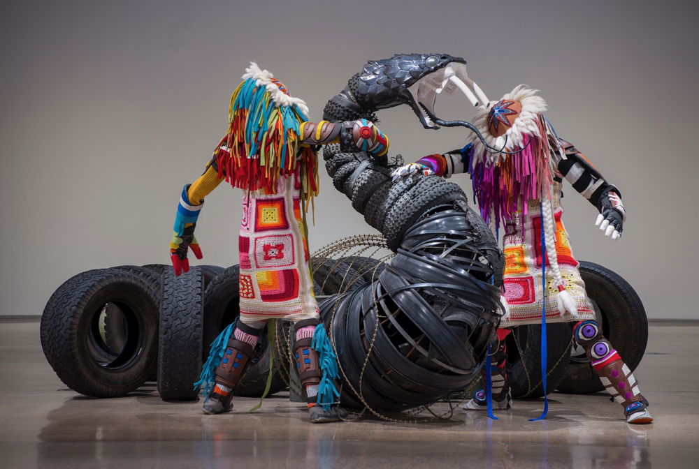An installation in a white walled museum. Two bright colored life-size humanoid figures with ceramic faced regalia engage with a massive ceramic headed snake form whose body is created by detritus of industry.