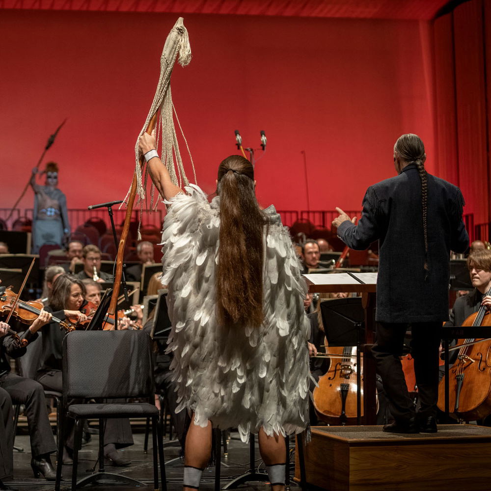 Jerod Tate, a Chickasaw man with long brown hair, stands facing away from the camera towards a symphony orchestra. Tate is dressed in ancient Chickasaw regalia, a tunic with gray feathers, and holds a staff high in his left hand. Towards the back left of the photo, a Chickasaw actor also in regalia is seen holding a staff high in their hand, facing Tate.