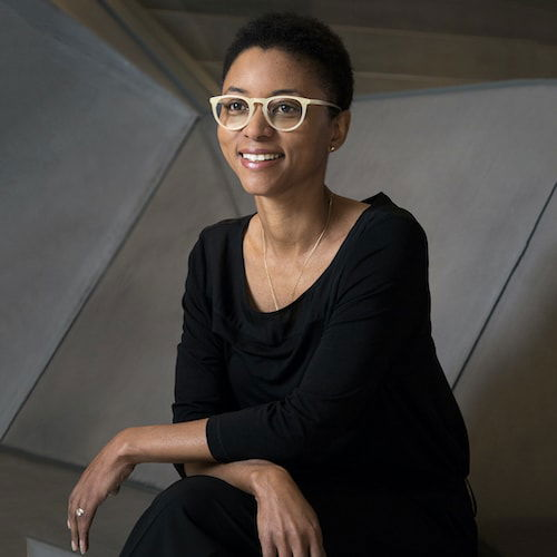 Portrait of an African American woman with short hair and brown skin. She sits leaning slightly forward with her arms crossed across her knees. She is dressed in all-black and is wearing yellow-rimmed glasses. She sits in front of a geometric volume in the background.