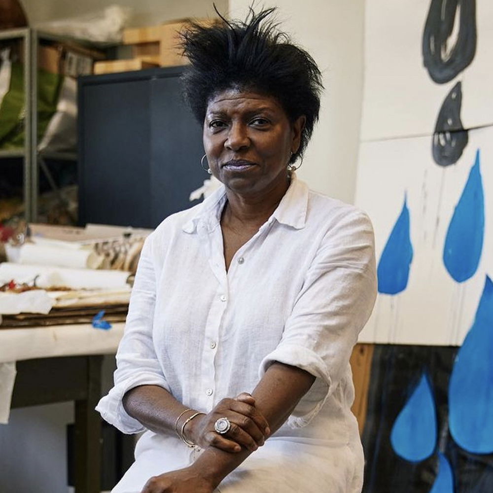 A woman poses on a stool in a studio space wearing a long white dress. Works in progress are laid out on a table behind her. A large work depicting rain droplets in black and blue hangs on the back wall.
